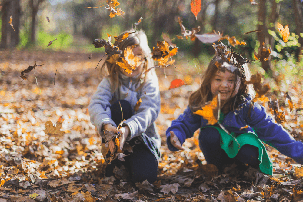 Due bambine esplorano e giocano con foglie autunnali durante un'esperienza con materiali naturali all'aperto.
